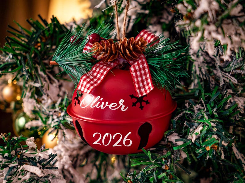 May include: A red metal jingle bell ornament featuring the name Oliver and the year 2026 in white script. The top is decorated with a red and white checkered ribbon, two small pinecones, green pine needles, and red berries.