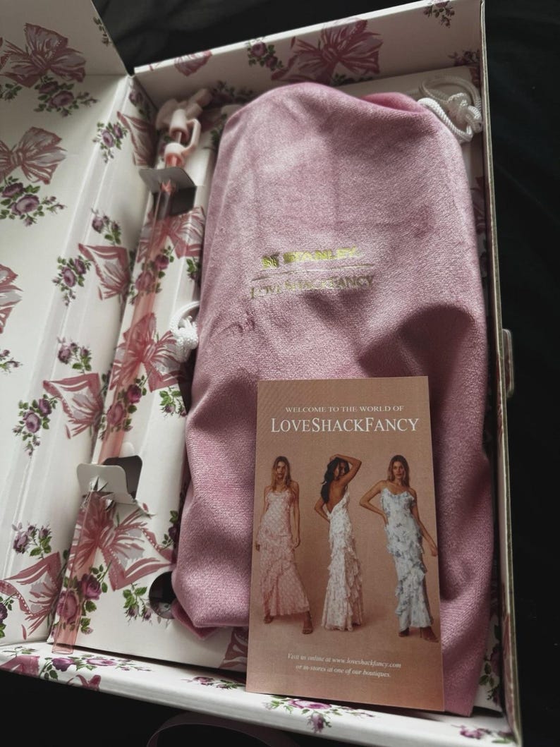 May include: A pink velvet drawstring pouch with the gold embroidered text STANLEY LOVESHACKFANCY inside a floral patterned box. A rectangular card with the text WELCOME TO THE WORLD OF LOVESHACKFANCY and images of three dresses rests on the pouch.