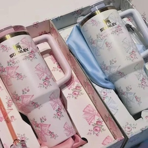 May include: Two Stanley tumblers with handles, one in light pink and one in white, both featuring a floral and bow pattern. The tumblers are branded with the Stanley logo and the text LOVESHACKFANCY, presented in a matching floral gift box.