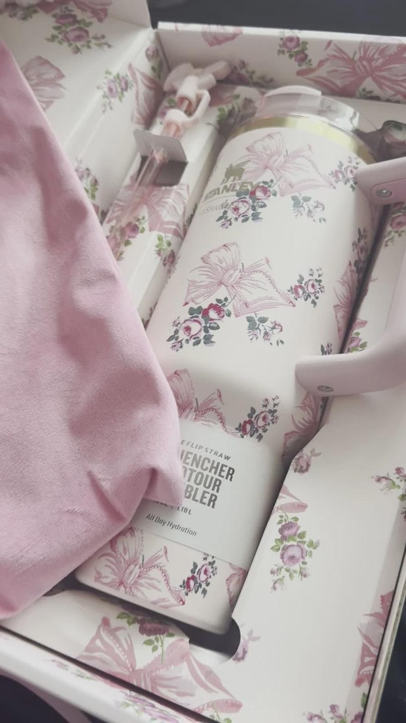 May include: A white Stanley Quencher H2.0 FlowState tumbler with a pink floral and ribbon pattern, featuring a matching pink handle and straw, packaged in a decorative box with a pink fabric item and text reading Stanley, Quencher, and 1.18 L.
