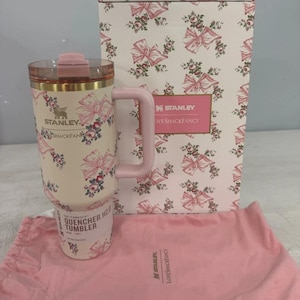 May include: A cream-colored Stanley Quencher H2.0 tumbler featuring a pink bow and floral pattern, a pink handle, and a clear lid. It is accompanied by a matching patterned box and a pink fabric drawstring bag, both branded with Stanley and LoveShackFancy.