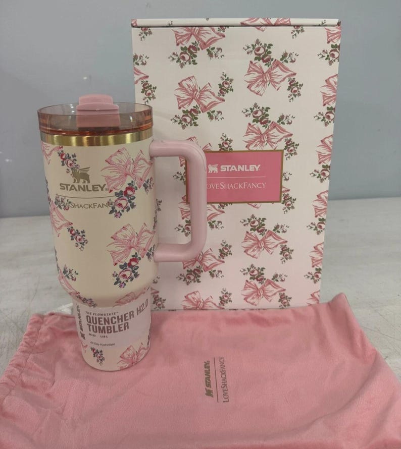 May include: A cream-colored Stanley Quencher H2.0 tumbler featuring a pink bow and floral pattern, a pink handle, and a clear lid. It is accompanied by a matching patterned box and a pink fabric drawstring bag, both branded with Stanley and LoveShackFancy.