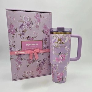 May include: A lavender Stanley tumbler with a floral pattern and a matching handle, positioned next to a rectangular gift box featuring the same floral design, a pink ribbon, and the text Stanley LoveShackFancy printed on the front of both items.