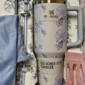 May include: A cream-colored Stanley LoveShackFancy 40 oz FlowState Quencher H2.0 tumbler featuring a purple floral and bow pattern, a matching straw with a bow topper, a blue drawstring pouch, and a promotional card in a branded gift box.