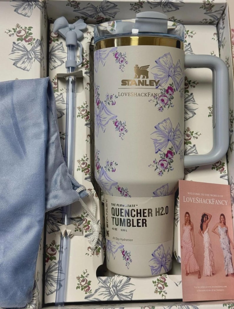 May include: A cream-colored Stanley LoveShackFancy 40 oz FlowState Quencher H2.0 tumbler featuring a purple floral and bow pattern, a matching straw with a bow topper, a blue drawstring pouch, and a promotional card in a branded gift box.