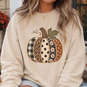 May include: A sand-colored crewneck sweatshirt featuring a large central pumpkin graphic. The pumpkin is composed of vertical segments with varied patterns including black and white buffalo plaid, leopard print, black polka dots, and floral prints.