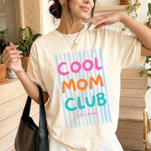 Colorful retro typography ‘Cool Mom Club’ design with pink, orange, and teal lettering over light blue vertical stripes, featuring small ‘EST 2026’ text on a Cream Comfort Colors  t-shirt.