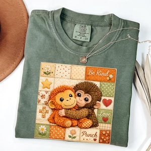 May include: Sage green Comfort Colors t-shirt with a patch featuring two hugging monkey plushies. The patch includes the words "Be Kind", "Love", and "Punch". A silver necklace with a pendant is draped on the shirt.