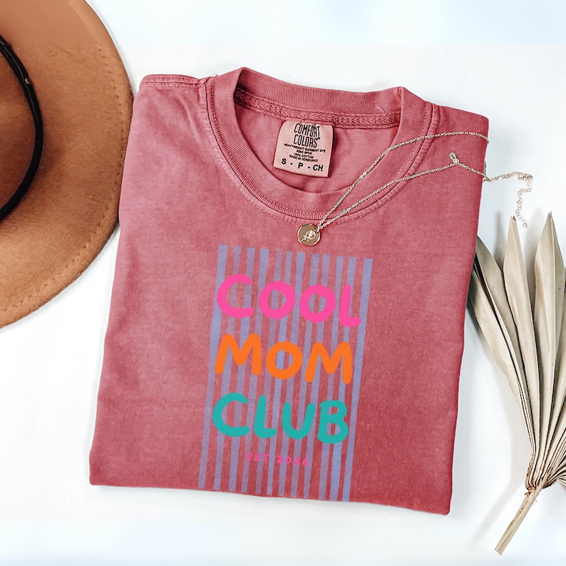 Colorful retro typography ‘Cool Mom Club’ design with pink, orange, and teal lettering over light blue vertical stripes, featuring small ‘EST 2026’ text on a coral Comfort Colors t-shirt.
