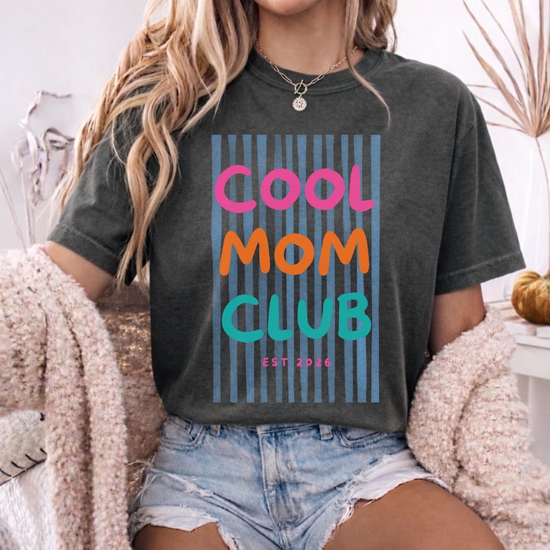 Colorful retro typography ‘Cool Mom Club’ design with pink, orange, and teal lettering over light blue vertical stripes, featuring small ‘EST 2026’ text on a gray Comfort Colors t-shirt.