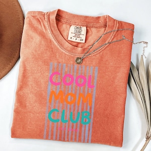 Colorful retro typography ‘Cool Mom Club’ design with pink, orange, and teal lettering over light blue vertical stripes, featuring small ‘EST 2026’ text on a orange Comfort Colors t-shirt.