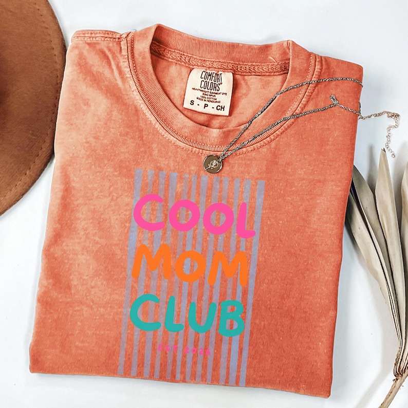 Colorful retro typography ‘Cool Mom Club’ design with pink, orange, and teal lettering over light blue vertical stripes, featuring small ‘EST 2026’ text on a orange Comfort Colors t-shirt.