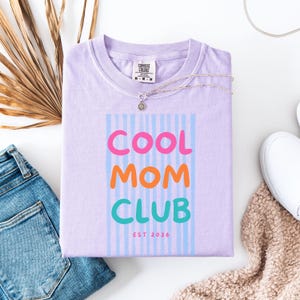 Colorful retro typography ‘Cool Mom Club’ design with pink, orange, and teal lettering over light blue vertical stripes, featuring small ‘EST 2026’ text on a purple Comfort Colors t-shirt.
