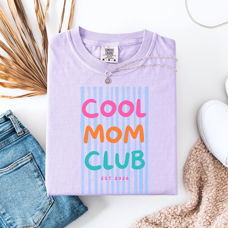 Colorful retro typography ‘Cool Mom Club’ design with pink, orange, and teal lettering over light blue vertical stripes, featuring small ‘EST 2026’ text on a purple Comfort Colors t-shirt.