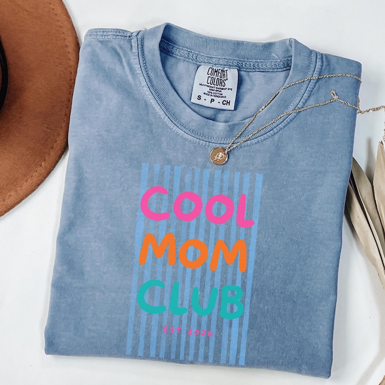 Colorful retro typography ‘Cool Mom Club’ design with pink, orange, and teal lettering over light blue vertical stripes, featuring small ‘EST 2026’ text on a blue Comfort Colors t-shirt.