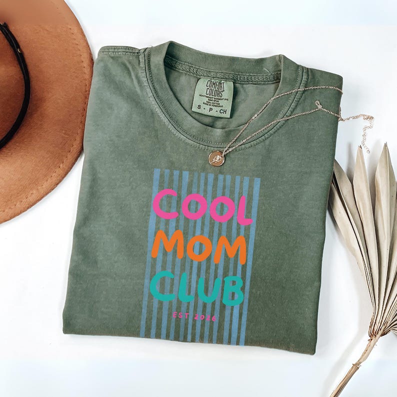 Colorful retro typography ‘Cool Mom Club’ design with pink, orange, and teal lettering over light blue vertical stripes, featuring small ‘EST 2026’ text on a grey Comfort Colors t-shirt.
