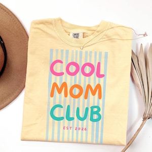 Colorful retro typography ‘Cool Mom Club’ design with pink, orange, and teal lettering over light blue vertical stripes, featuring small ‘EST 2026’ text on a yellow Comfort Colors t-shirt.