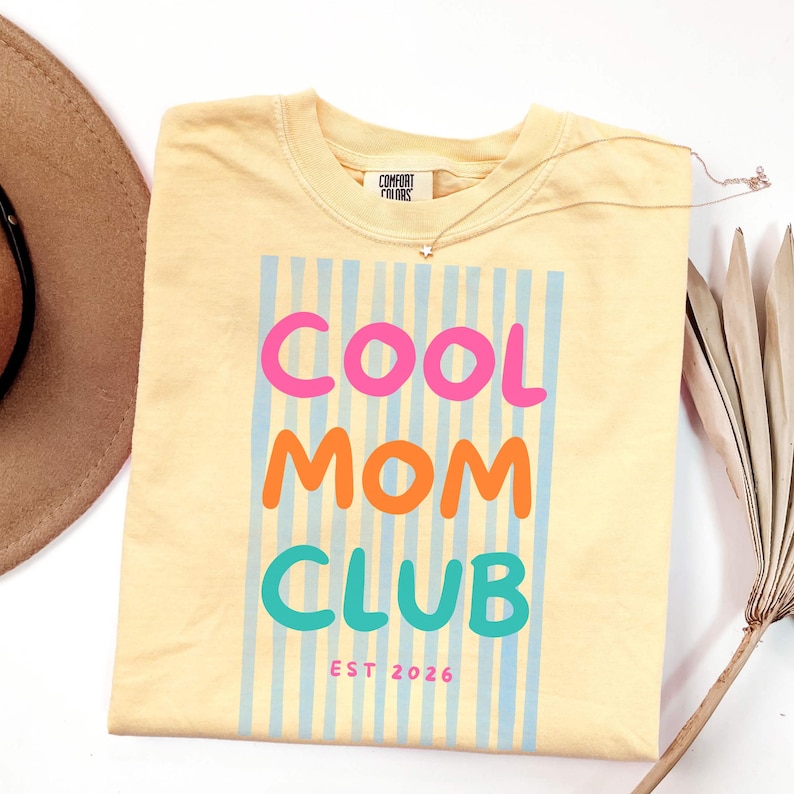 Colorful retro typography ‘Cool Mom Club’ design with pink, orange, and teal lettering over light blue vertical stripes, featuring small ‘EST 2026’ text on a yellow Comfort Colors t-shirt.