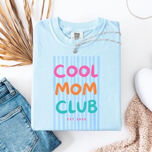 Colorful retro typography ‘Cool Mom Club’ design with pink, orange, and teal lettering over light blue vertical stripes, featuring small ‘EST 2026’ text on a blue Comfort Colors t-shirt.