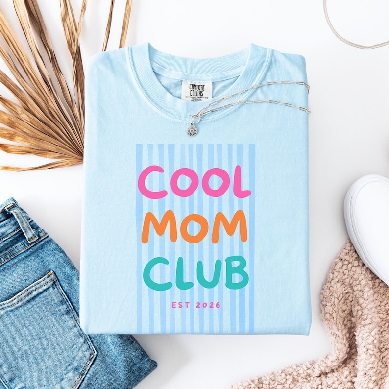 Colorful retro typography ‘Cool Mom Club’ design with pink, orange, and teal lettering over light blue vertical stripes, featuring small ‘EST 2026’ text on a blue Comfort Colors t-shirt.