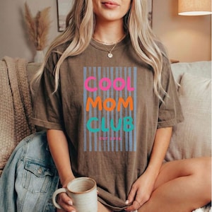 Colorful retro typography ‘Cool Mom Club’ design with pink, orange, and teal lettering over light blue vertical stripes, featuring small ‘EST 2026’ text on a brown  Comfort Colors t-shirt.