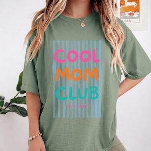 Colorful retro typography ‘Cool Mom Club’ design with pink, orange, and teal lettering over light blue vertical stripes, featuring small ‘EST 2026’ text on a green Comfort Colors t-shirt.