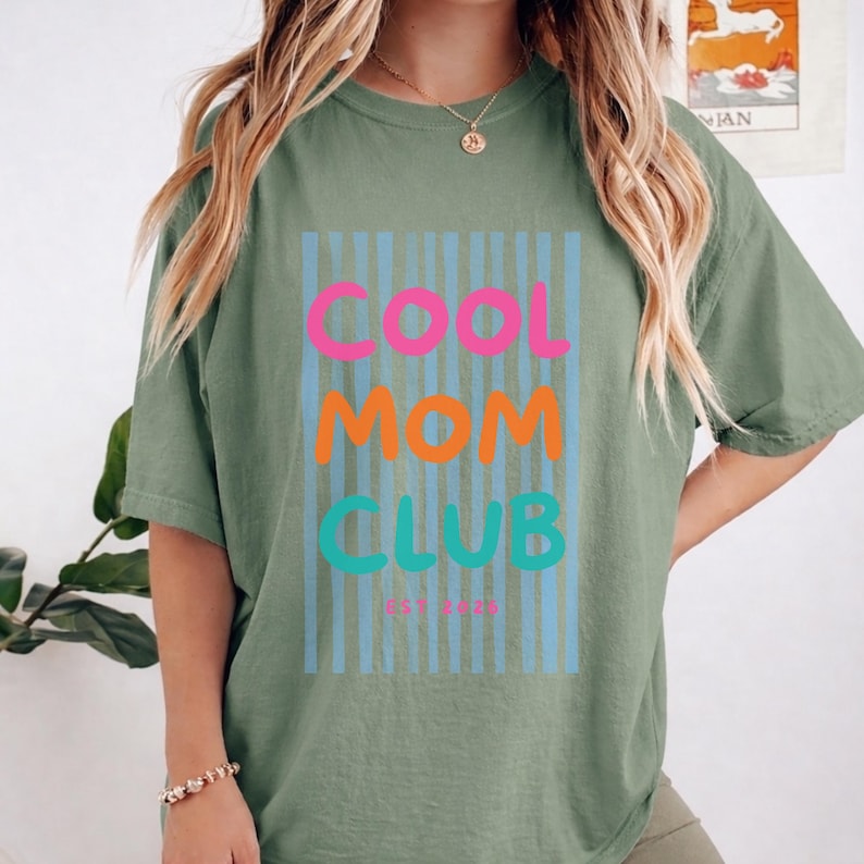 Colorful retro typography ‘Cool Mom Club’ design with pink, orange, and teal lettering over light blue vertical stripes, featuring small ‘EST 2026’ text on a green Comfort Colors t-shirt.
