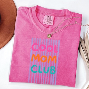 Colorful retro typography ‘Cool Mom Club’ design with pink, orange, and teal lettering over light blue vertical stripes, featuring small ‘EST 2026’ text on a pink Comfort Colors t-shirt.