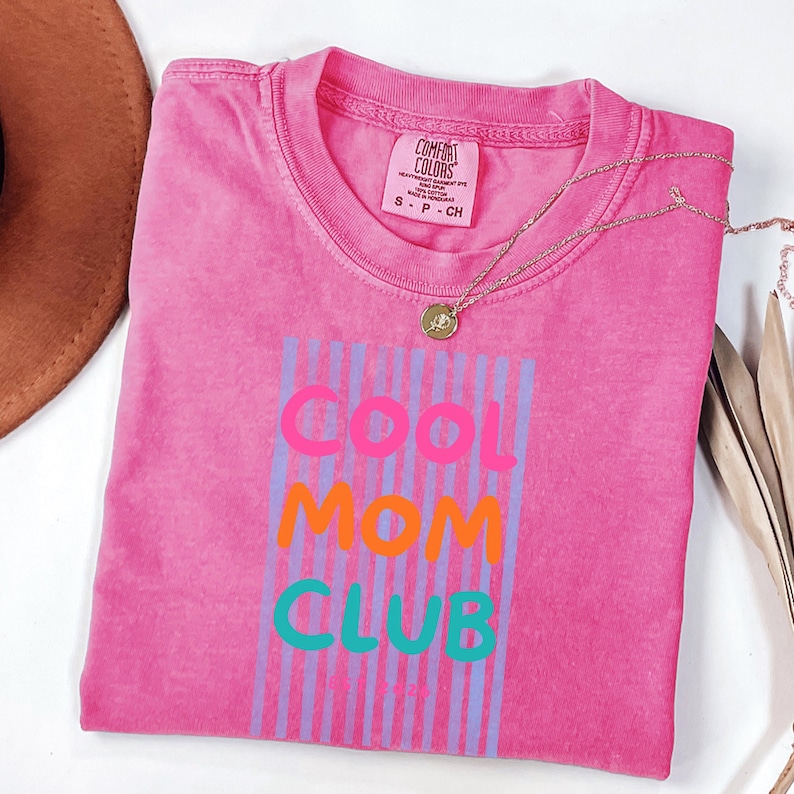 Colorful retro typography ‘Cool Mom Club’ design with pink, orange, and teal lettering over light blue vertical stripes, featuring small ‘EST 2026’ text on a pink Comfort Colors t-shirt.