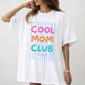Colorful retro typography ‘Cool Mom Club’ design with pink, orange, and teal lettering over light blue vertical stripes, featuring small ‘EST 2026’ text on a white Comfort Colors t-shirt.