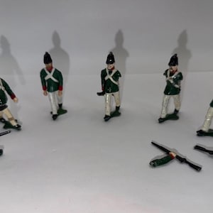 May include: A set of six miniature toy soldiers painted in green jackets, white trousers, and black hats. One soldier holds a rifle, while two separate rifles lie on the surface. The figures are standing in a loose formation on a plain white background.