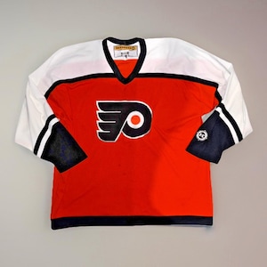 May include: A red and white hockey jersey with the Philadelphia Flyers logo on the front. The jersey has black accents on the sleeves and collar. The sleeves are white with black stripes. The jersey has a V-neck collar.