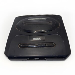 Może przedstawiać: Czarna konsola do gier wideo Sega Genesis z gniazdem na kartridże ładowanym od góry, wyraźnym logo Sega na przednim panelu oraz przyciskiem zasilania z poziomą lampką kontrolną. Konsola ma smukłą, zakrzywioną obudowę z otworami wentylacyjnymi.