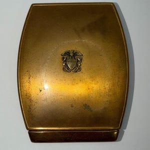 May include: A rectangular brass compact case with rounded sides and a polished finish. The center of the lid features a raised emblem of an eagle perched above a shield with crossed anchors. The metal surface shows light surface scratches.