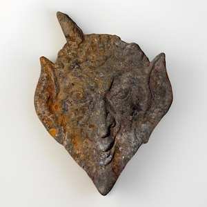 Könnte beinhalten: Eine verwitterte Metallskulptur eines Gesichts mit spitzen Ohren und einem einzelnen Horn. Die rautenförmige Skulptur weist Brauntöne, Grau und Orange auf, was auf Rost und Alter hindeutet. Das Gesicht hat detaillierte Merkmale.