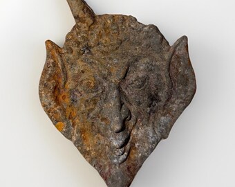 May include: A weathered, textured metal sculpture of a face with pointed ears and a single horn. The sculpture has a diamond shape and is colored in shades of brown, gray, and orange, suggesting rust and age. The face has detailed features.