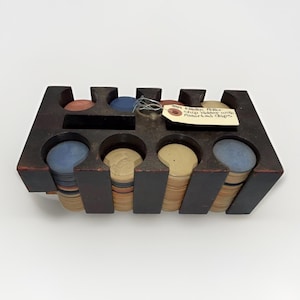 May include: A dark brown wooden poker chip holder containing stacks of assorted colored chips. A beige paper tag is attached to the holder with the handwritten text: "Old Wooden Poker Chip Holder with Assorted Chips". The holder has eight slots.
