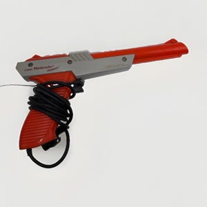 Puede incluir: Una pistola controladora Nintendo Zapper de color rojo y gris con un cañón largo y un cable negro enrollado alrededor del mango. El lateral del dispositivo presenta el logotipo de Nintendo Zapper impreso en negro sobre la carcasa gris.