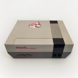 Könnte beinhalten: Eine graue und schwarze Nintendo Entertainment System Videospielkonsole mit einem roten Nintendo Entertainment System Logo auf der Vorderseite und einem kleinen Aufkleber einer Figur mit roter Mütze und Schnurrbart auf der Oberseite.
