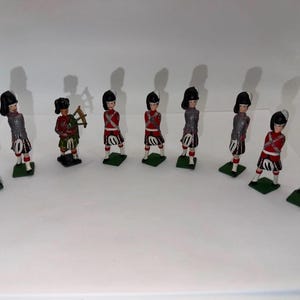 Puede incluir: Un conjunto de nueve figuras de soldados en miniatura sobre pequeñas bases rectangulares verdes. Las figuras llevan altos gorros negros de piel de oso, chaquetas grises o rojas y faldas escocesas. Una figura sostiene una gaita y otras posan.