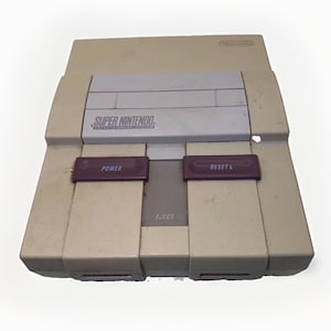 Puede incluir: Una consola de videojuegos Super Nintendo Entertainment System de color beige con una cubierta gris para la ranura de cartuchos, dos botones rectangulares morados etiquetados como Power y Reset, y un botón Eject gris central. La carcasa muestra desgaste.