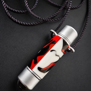 May include: A cylindrical pendant featuring a red, white, and black marbled pattern, capped with silver-toned metal ends. The pendant is attached to a dark, twisted cord necklace that loops around the top, creating a secure hanging mechanism.