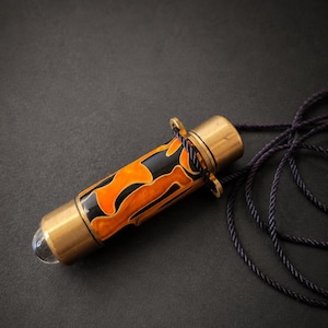 May include: A cylindrical pendant featuring a marbled orange and black resin body encased in brass caps, with a clear glass dome at one end and a dark purple twisted cord attached to a brass loop for wearing as a necklace.