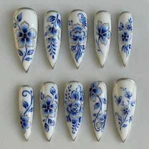May include: A set of ten white and blue floral design press-on nails. Each nail features a different blue floral pattern with silver metallic accents. The nails are almond-shaped and have a glossy finish, perfect for a unique manicure.