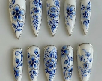 May include: A set of ten white and blue floral design press-on nails. Each nail features a different blue floral pattern with silver metallic accents. The nails are almond-shaped and have a glossy finish, perfect for a unique manicure.