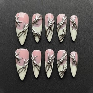 May include: Set of ten artificial nails. Each nail is almond-shaped with a white tip and a pink base. Silver-colored barbed wire designs are affixed to each nail, adding a unique, edgy aesthetic. The nails are arranged on a dark surface.
