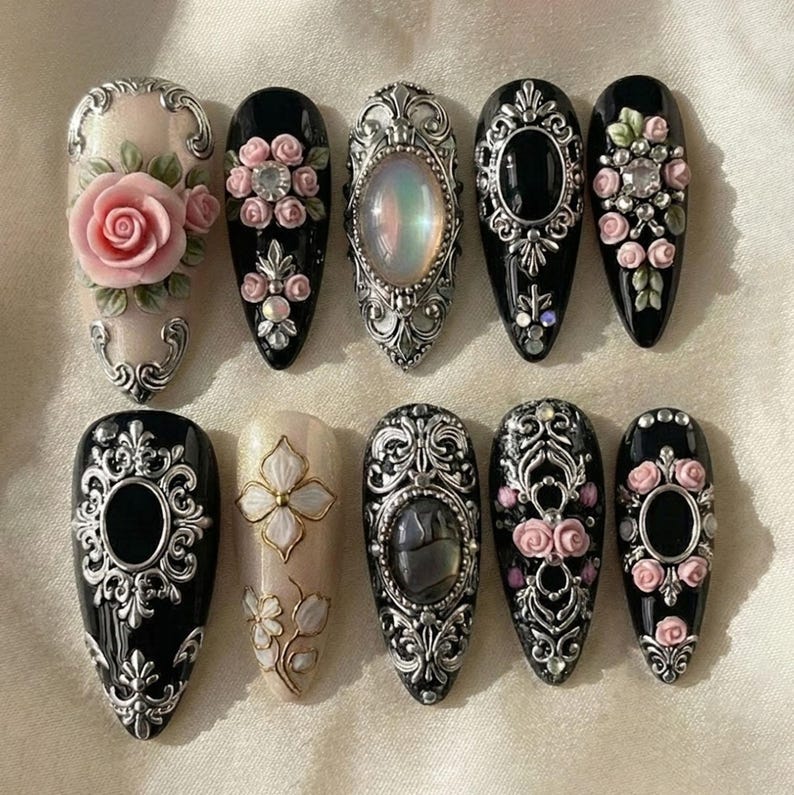 May include: A collection of ten artificial nails with ornate designs. The nails feature a variety of colors, including black, cream, and silver, and are adorned with roses, gems, and intricate patterns. Some nails have oval or gemstone accents.