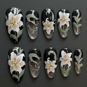 K&ouml;nnte beinhalten: Ein Set aus zehn ovalen Press-On-N&auml;geln. Jeder Nagel hat eine schwarze Basis mit einem handgemalten wei&szlig;en Lilien-Design, gr&uuml;nen Bl&auml;ttern und silbernen Glitzerdetails. Die N&auml;gel sind in verschiedenen Gr&ouml;&szlig;en erh&auml;ltlich.
