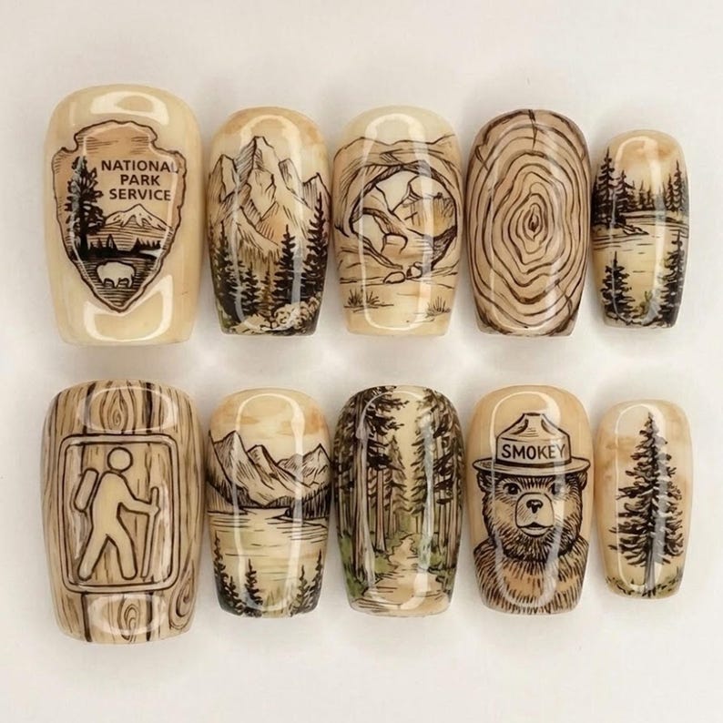 May include: A set of ten beige-colored, oval-shaped nail art designs. Each nail features a unique hand-drawn illustration of nature scenes, including mountains, trees, and a bear wearing a hat. Some nails display the text "NATIONAL PARK SERVICE" and "SMOKEY".