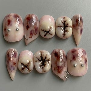 Könnte beinhalten: Ein Set aus zehn künstlichen Fingernägeln mit einem einzigartigen Design. Die Nägel haben eine hellrosa Basis mit marmorierten Mustern in Burgunderrot und Braun. Einige Nägel sind wie Zähne geformt, mit detaillierten Zahnwurzel- und Zahnschmelzdesigns. Silberne Nieten und Ringe sind hinzugefügt.